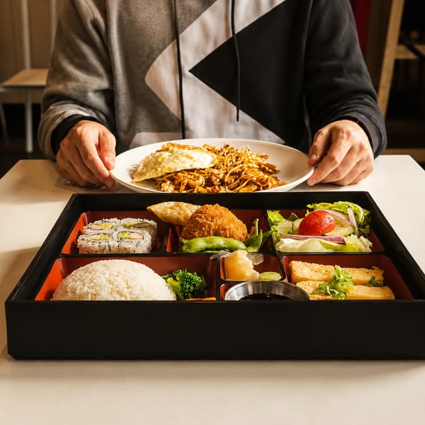 A person holding a plate of noodles and egg, with a bento box of sushi, rice, and fried chicken.
