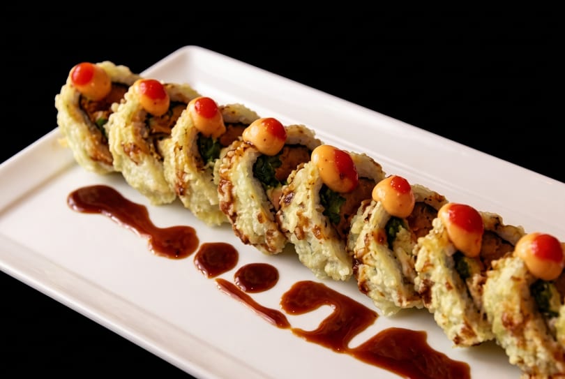 Crispy fried sushi rolls with spicy mayo and red dots, arranged on a white rectangular plate.