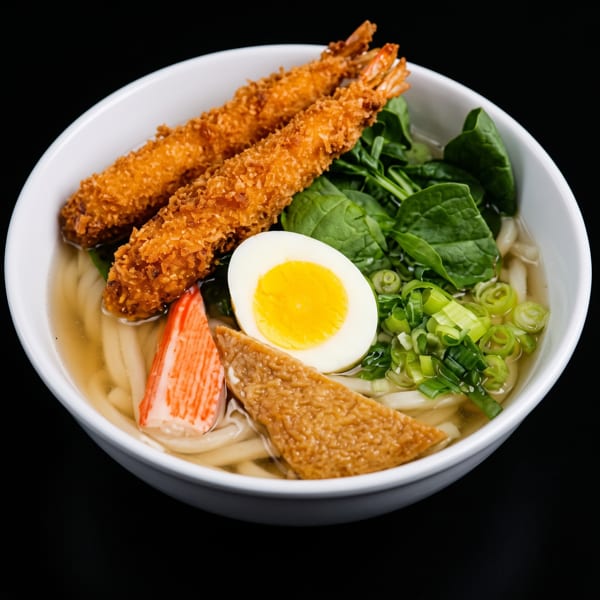 Udon noodle soup with tempura shrimp, spinach, egg, crab stick, and scallions in a white bowl.