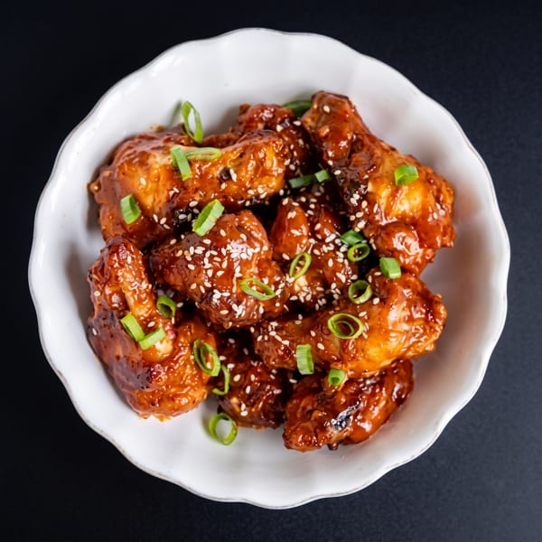 A white bowl filled with sticky, glazed chicken wings, garnished with sesame seeds and chopped green onions.