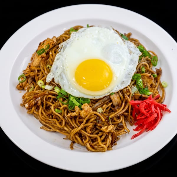 Yakisoba noodles with chicken, green onions, red pickled ginger, and a fried egg on a white plate.